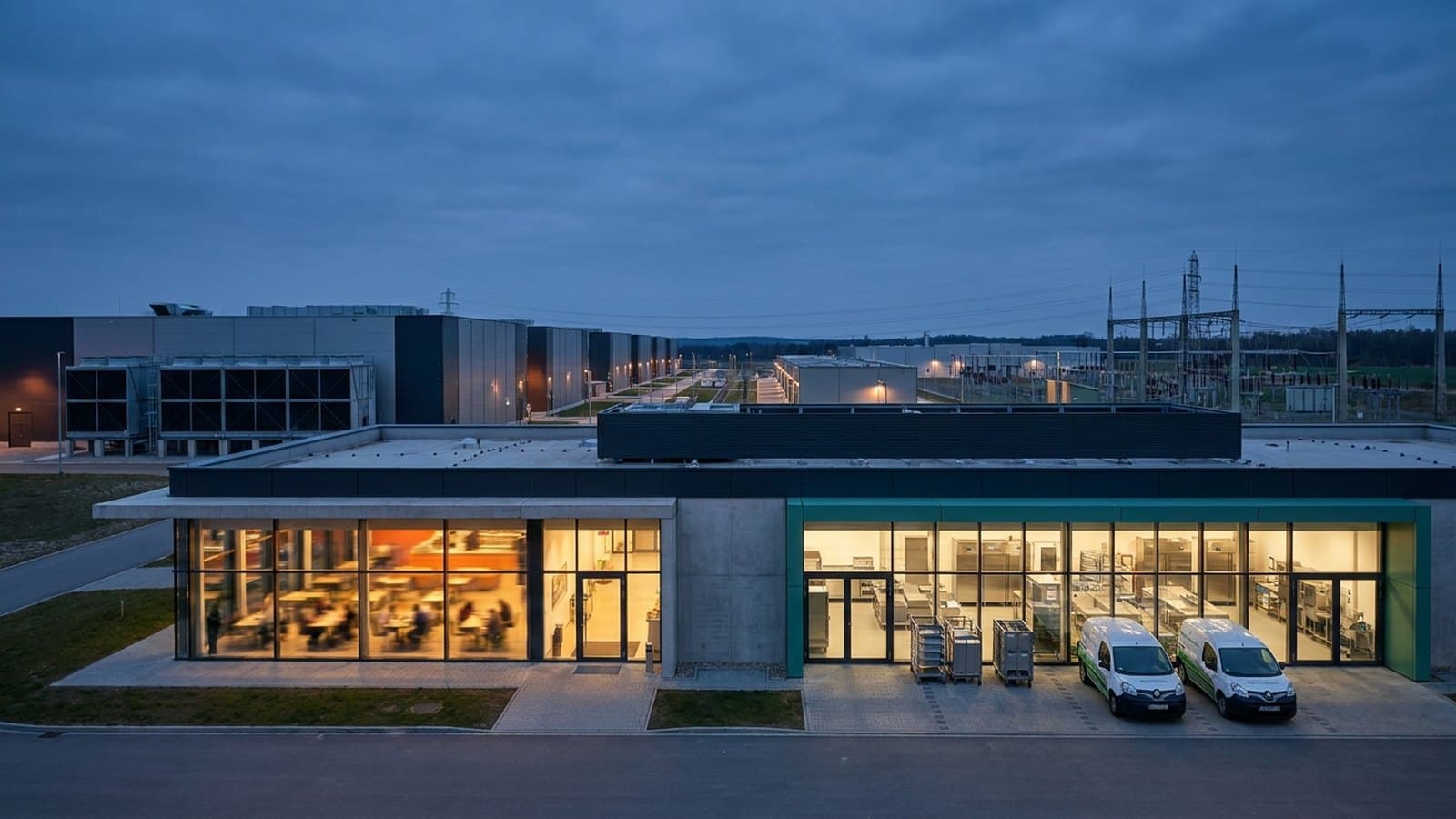 A large modern industrial catering and dining facility supporting a hyperscale data center campus at dusk, with the low server-hall buildings and power infrastructure visible on the horizon.