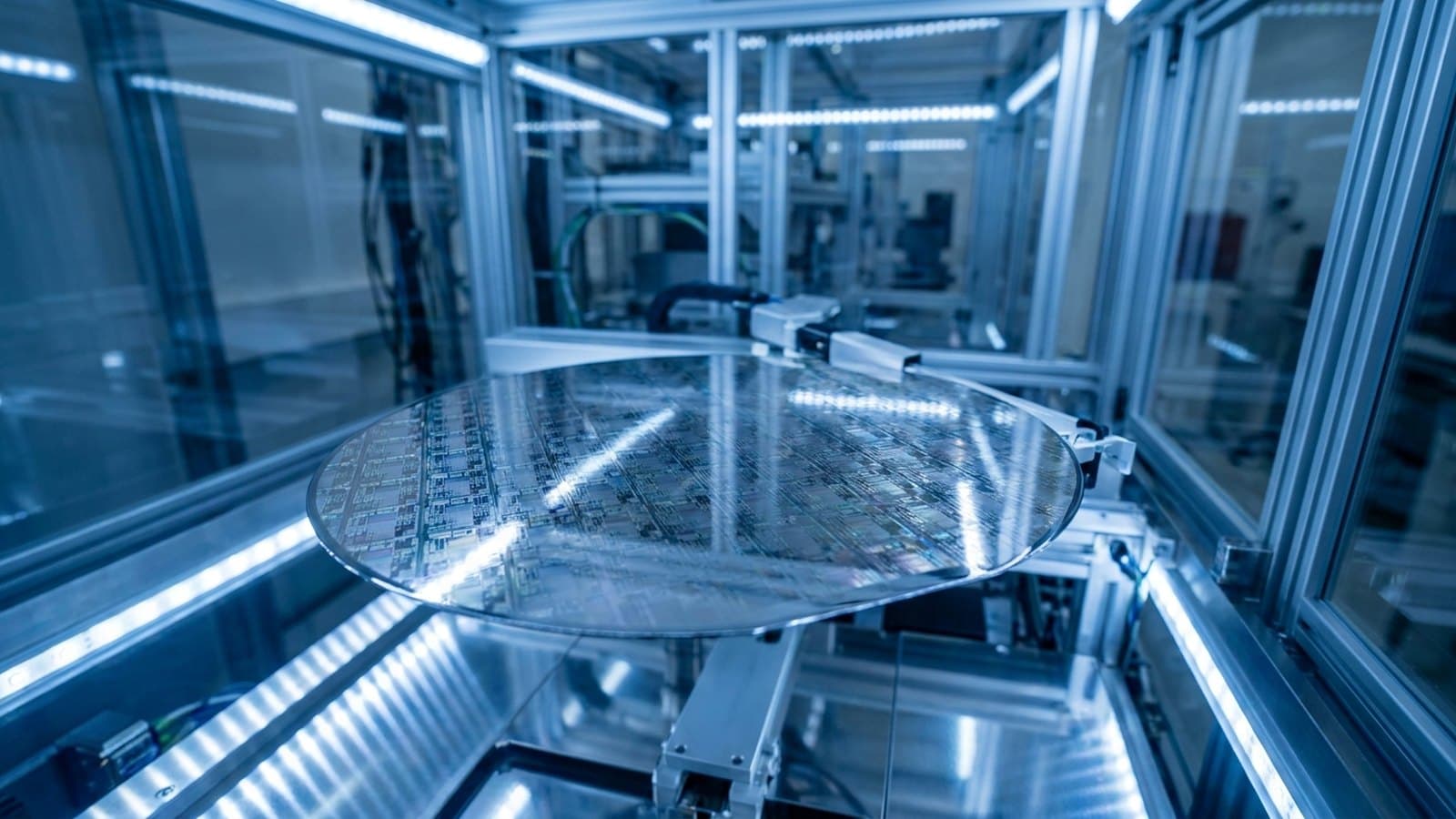 Extreme close-up of a polished silicon wafer covered in mirror-like microchip circuitry, held on a robotic wafer-handling arm inside an advanced semiconductor fabrication tool under cool blue clean-room lighting, representing Intel's foundry-manufacturing turnaround.