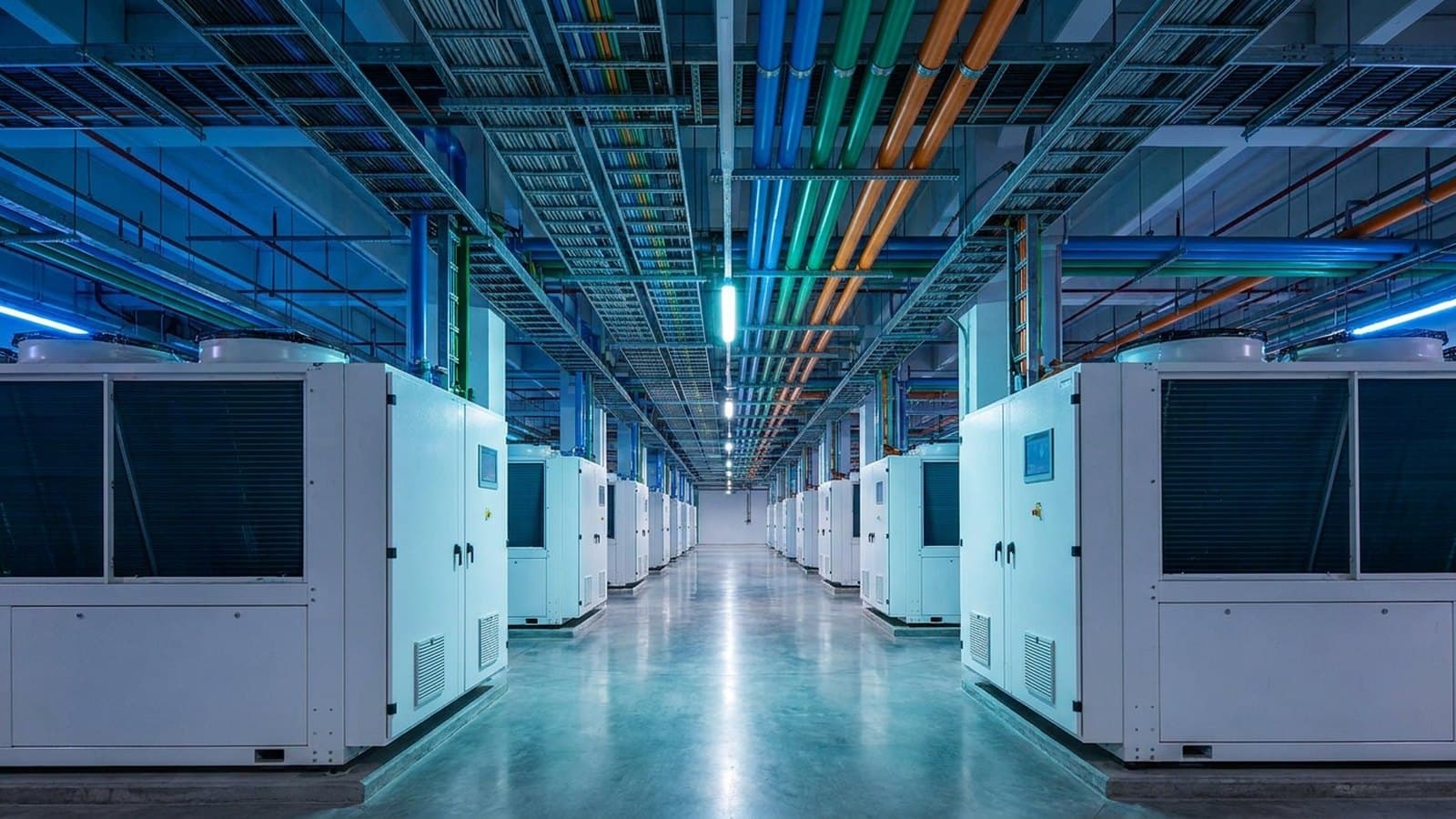 Wide view inside a modern data center's mechanical and electrical infrastructure — rows of cooling air-handling units, overhead cable trays and colored power and cooling piping along a service corridor under cool blue lighting — representing Legence's data-center engineering and services business.