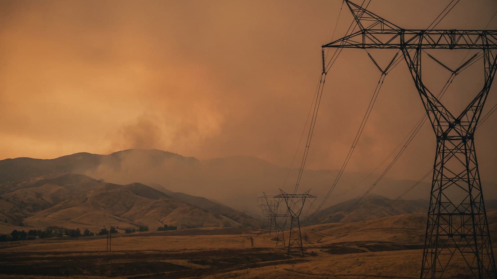 High-voltage electrical transmission towers and power lines silhouetted against a hazy orange, smoke-tinged California sky at dusk, evoking wildfire risk to the electric grid.
