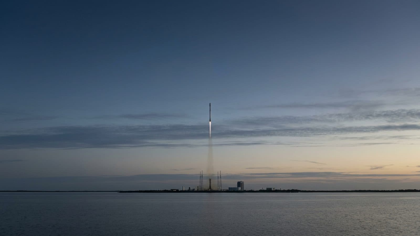 A Falcon-class rocket rising from a coastal launch pad at dawn against a wide banded sky, with the plume trailing across the horizon, illustrating SpaceX's 11.65% share-price gain on August 7, 2026