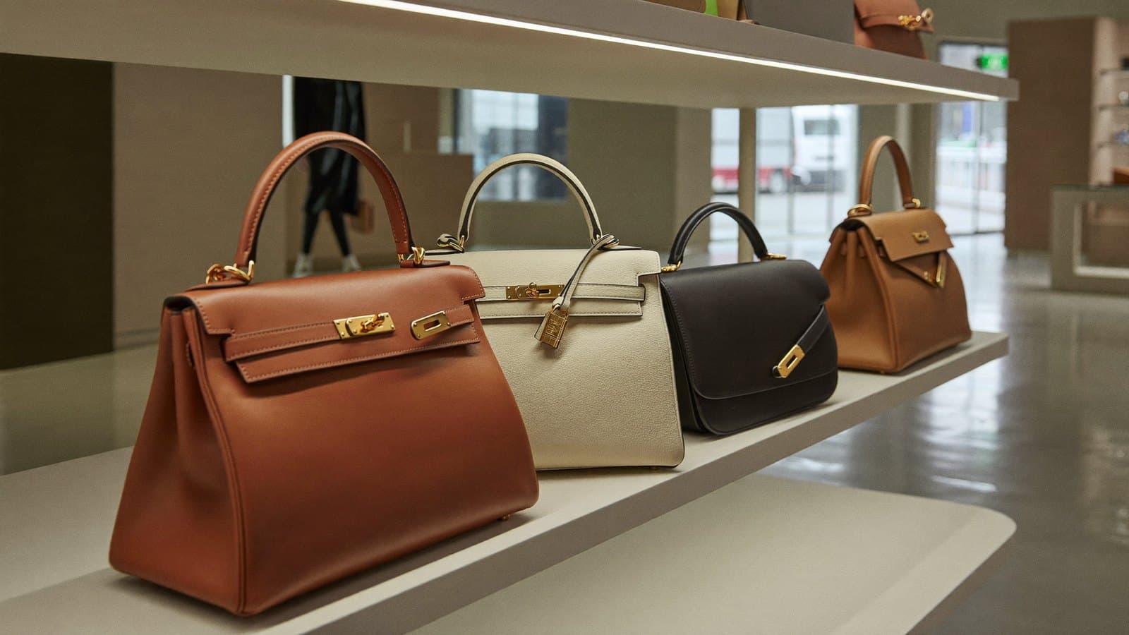 A row of structured leather handbags on a softly lit retail display shelf in a modern accessible-luxury store, shot at an angle with warm neutral tones and shallow depth of field.